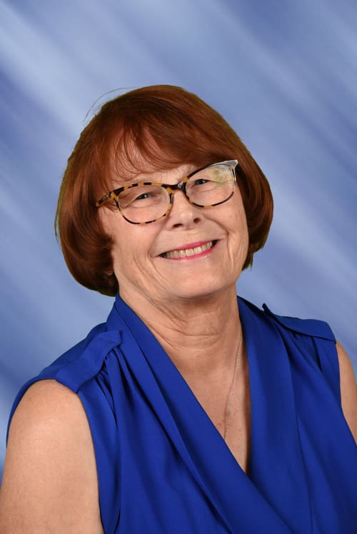Smiling older woman with glasses and short red hair in a blue sleeveless top.